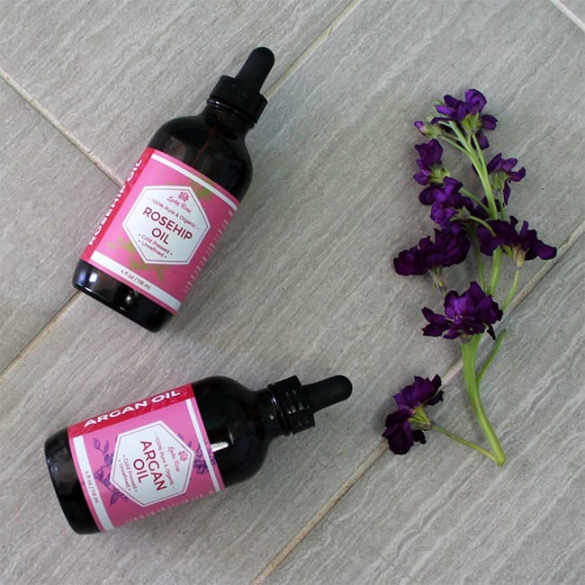 Custom-labeled bottles of rosehip and argan oil lying on the wood-look tiles next to the purple flower.