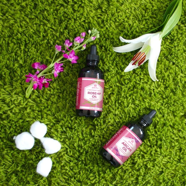 Custom-labeled bottles of rosehip and argan oil lying on the green carpet next to the purple and white flower.