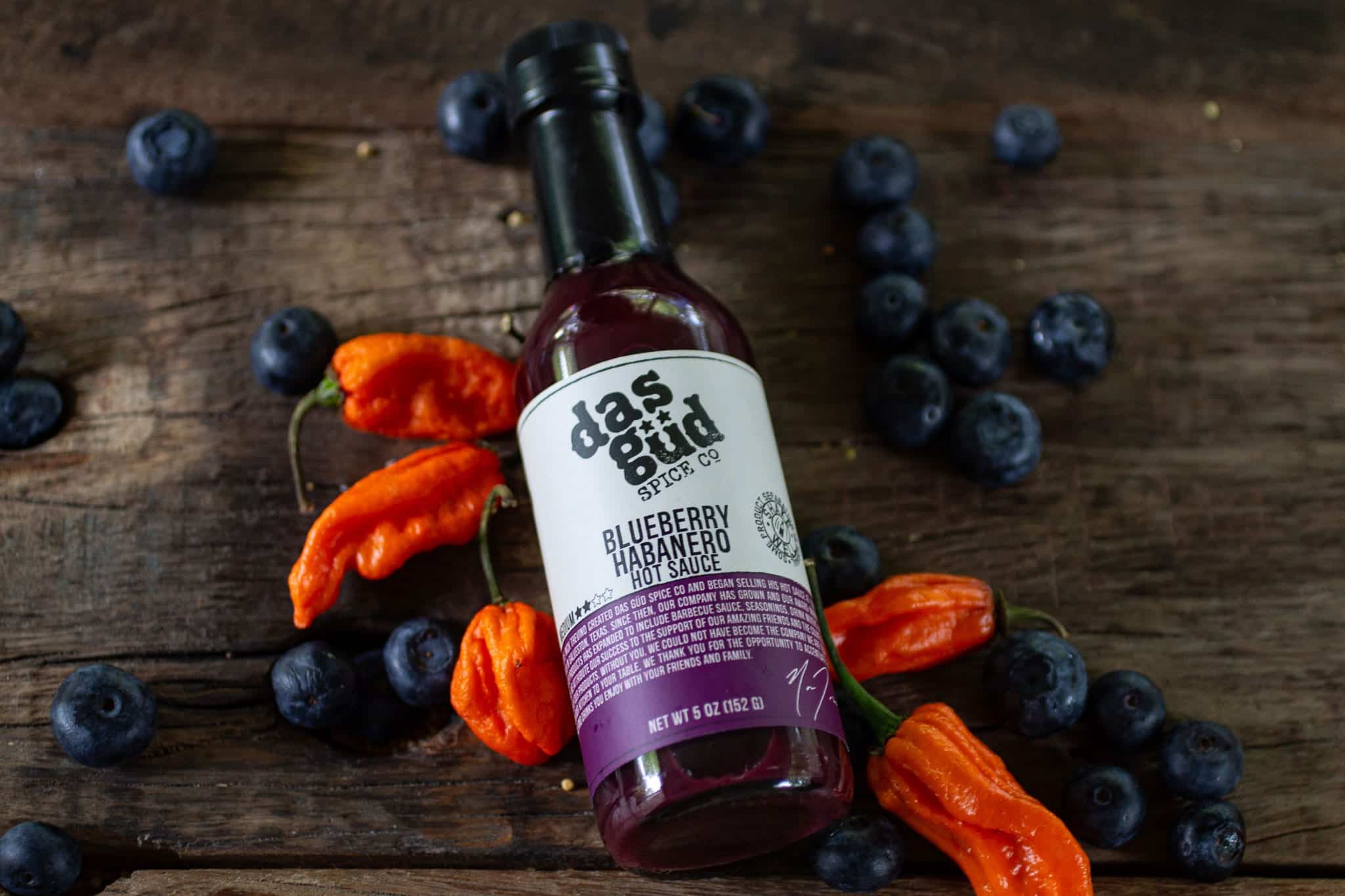 Custom-labeled bottle of Das Gut Hot Sauce, surrounded by fresh blueberries and habanero peppers on a wooden table.