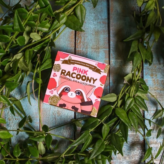 On a wooden surface surrounded by leaves, there is a cosmetic product custom label featuring a pinkish raccoon.