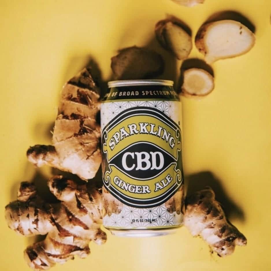 A custom labeled can of Sparkling CBD Ginger Ale lying on a yellow surface surrounded by ginger pieces.