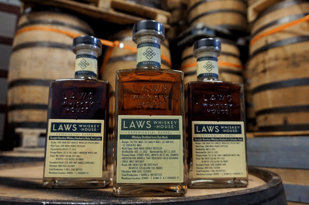 Three bottles of Laws Whiskey with embossed custom labels in a cellar surrounded by wooden barrels.