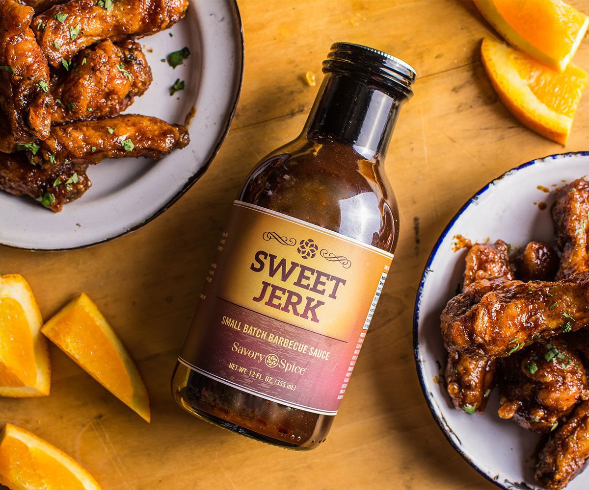 A custom-labeled bottle of Savory Spice Sweet Jerk surrounded by spiced chicken wings and orange slices on a wooden table.