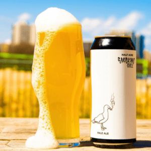 A Half Acre Smoking Gull custom labeled craft beer can sits next to an overfilled beer glass on a wooden surface.
