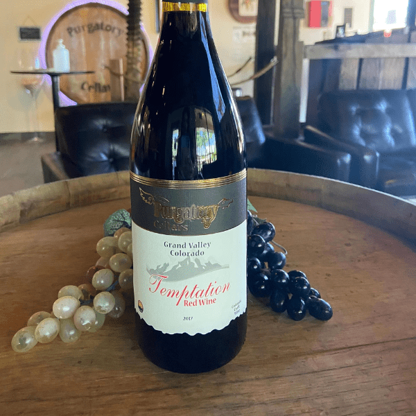 A wine bottle stands in front of black and white grapes on a wooden barrel.