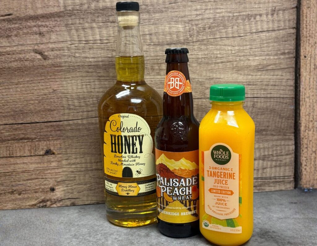 Three bottled drinks on a wooden backdrop: Colorado Honey bourbon whiskey (left), Palisade Peach Wheat beer (center), and Whole Foods Organic Tangerine Juice (right).